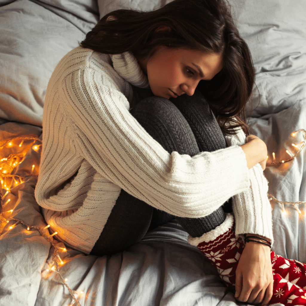 A woman looking depressed while sitting by holiday lights on her bed